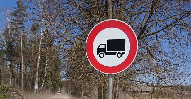 Apaļa ceļa zīme ar sarkanu apmali un melnu kravas automašīnas siluetu baltā fonā, kas norāda kravas automašīnu satiksmes aizliegumu. Fonā kaili koki, zilas debesis un grants ceļš.