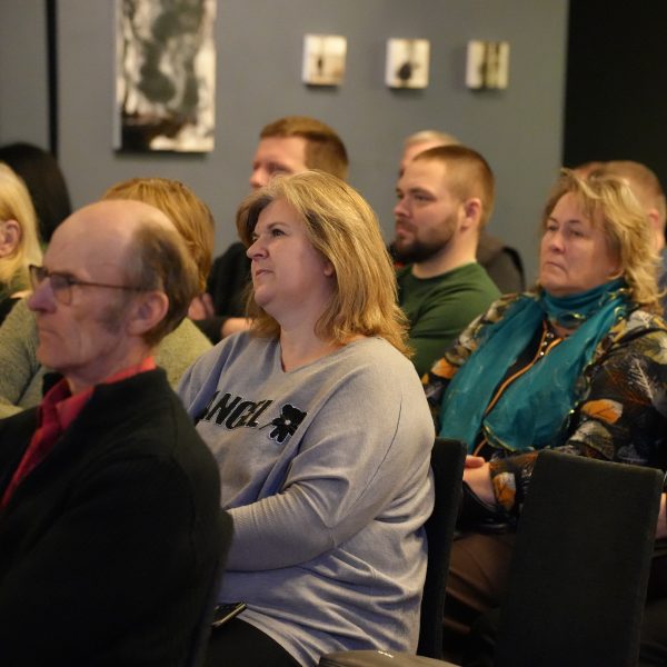 Daudzveidīga cilvēku grupa sēž auditorijā, skatoties uz priekšu.