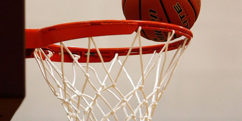 Tuvplānā redzama oranža basketbola bumba, kas atrodas uz sarkanas groza stīpas ar baltu tīklu. Fonā gaiša siena ar zilu svītru.