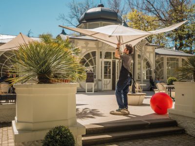 Vīrietis pelēkā kreklā un melnā priekšautā uzstāda lielu, gaišu saulessargu uz saulainas āra terases. Aiz viņa redzama eleganta ēka ar arkveida logiem un kupolu. Terasi rotā podos stādīti augi un liela sarkana piepūšamā bumba.