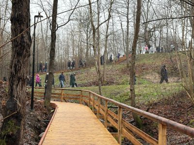 Koka laipa ar margām vijas cauri meža parkam, kur cilvēki pastaigājas pa takām un koka kāpnēm, kas ved augšup pa pakalnu.
