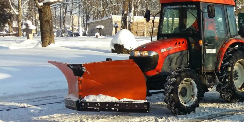 Sarkans traktors ar oranžu sniega lāpstu stāv uz sniegotas ceļa malas saulainā ziemas dienā.