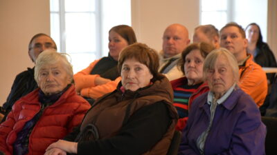 Cilvēku grupa, galvenokārt gados vecāki cilvēki, sēž iekštelpās, klausoties vai vērojot priekšā notiekošo. Priekšplānā redzamas trīs gados vecākas sievietes: viena ar baltiem matiem sarkanā jakā, otra ar brūniem matiem brūnā vestē, trešā ar sirmiem matiem violetā jakā.