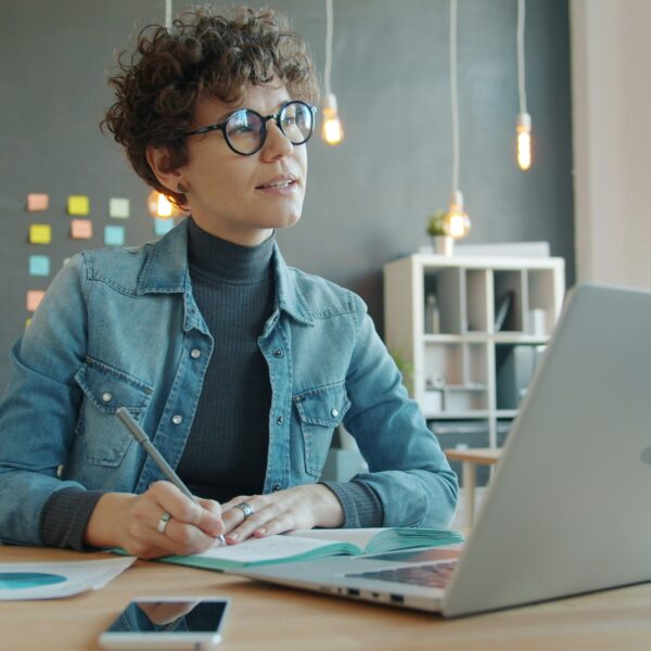 Pārdomās iegrimusi sieviete ar lokainiem matiem un brillēm sēž pie galda, rakstot piezīmju grāmatiņā ar pildspalvu, kamēr priekšā atvērts portatīvais dators modernā birojā.