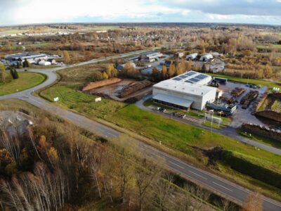Aerofotogrāfija attēlo rūpniecisku kompleksu ar lielu ēku, uz kuras fasādes redzams uzraksts "Enerģijas pētīnieki", un saules paneļiem uz jumta. Apkārt ēkai ir milzīgas baļķu kaudzes, ceļi, transportlīdzekļi un rudens koki.