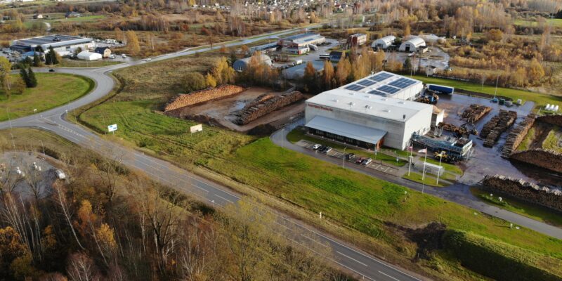 Aerofotogrāfija attēlo rūpniecisku kompleksu ar lielu ēku, uz kuras fasādes redzams uzraksts "Enerģijas pētīnieki", un saules paneļiem uz jumta. Apkārt ēkai ir milzīgas baļķu kaudzes, ceļi, transportlīdzekļi un rudens koki.